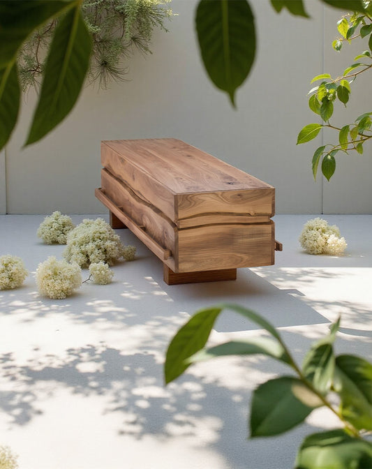 Massief iepenhouten uitvaartkist met zichtbare houtnerf, geplaatst in een serene, groene binnenplaats met schaduw en witte bloemen