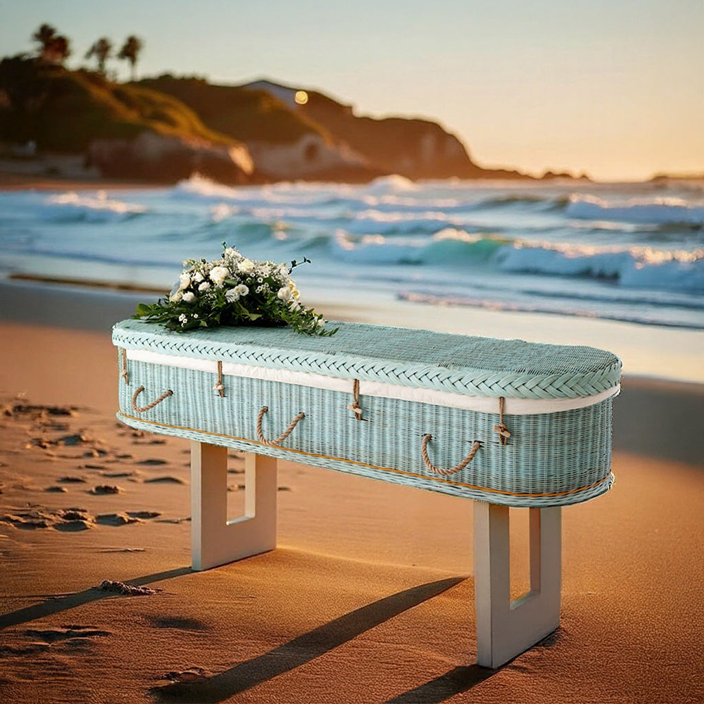Zachtblauwe rotan uitvaartmand Shore of Memories op het strand bij zonsondergang, met witte bloemen op het deksel.