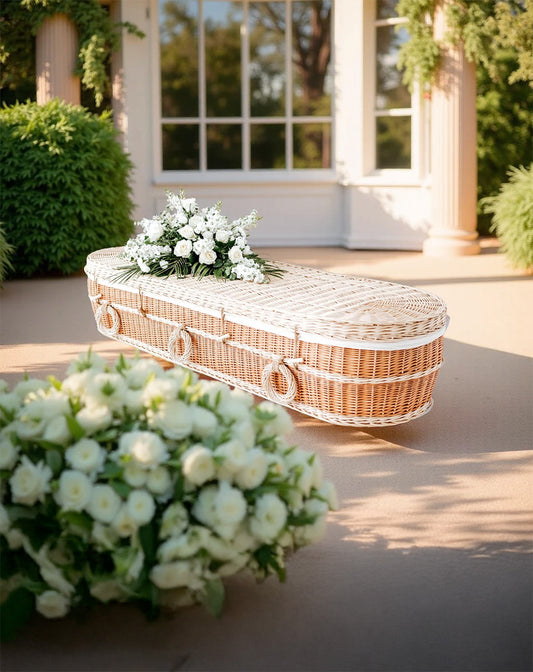 Lichtkleurige wilgenteen uitvaartmand Pure Elegance met witte bloemen, geplaatst in een klassieke tuinsetting.