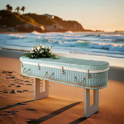 Zachtblauwe rotan uitvaartmand Shore of Memories op het strand bij zonsondergang, met witte bloemen op het deksel.