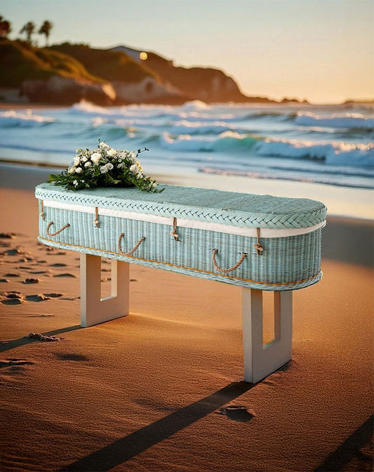 Zachtblauwe rotan uitvaartmand Shore of Memories op het strand bij zonsondergang, met witte bloemen op het deksel.