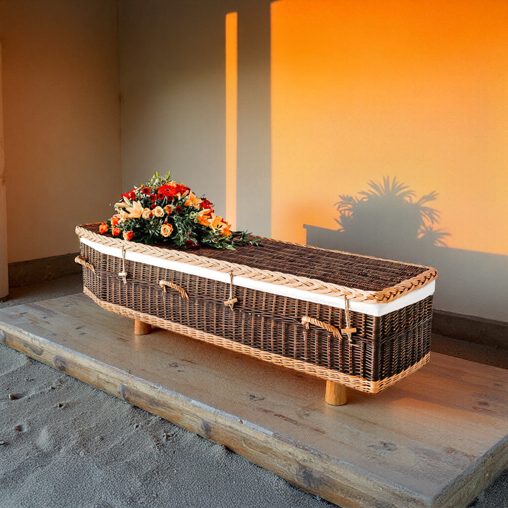 Donkerbruine wilgenteen uitvaartmand Sunset Farewell met lichte accenten en bloemen, in warme zonsondergangsgloed.