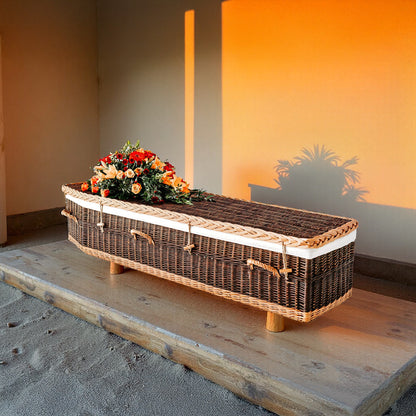 Donkerbruine wilgenteen uitvaartmand Sunset Farewell met lichte accenten en bloemen, in warme zonsondergangsgloed.